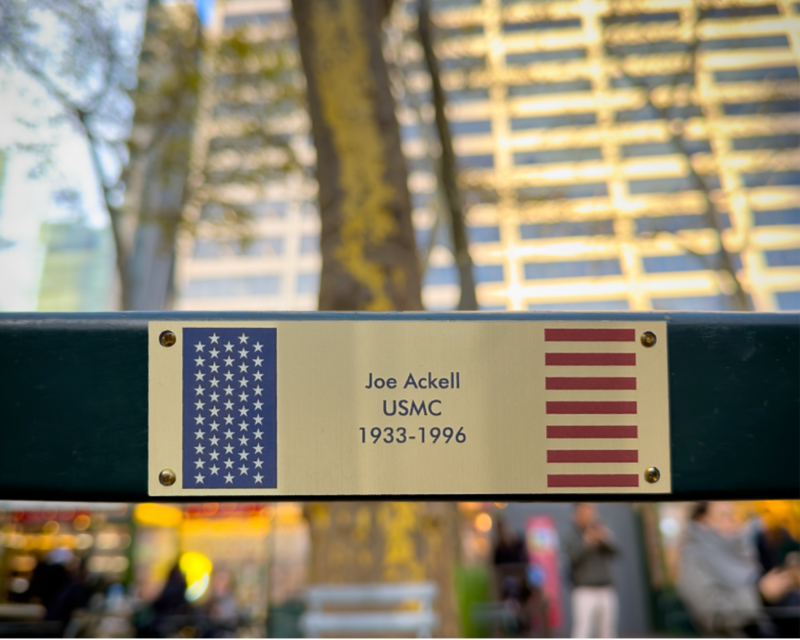 A close-up of a commemorative metal plaque mounted on a dark green horizontal bar. The plaque has a gold-toned background with a blue American flag on the left, red stripes on the right, and the text 'Joe Ackell USMC 1933-1996'.